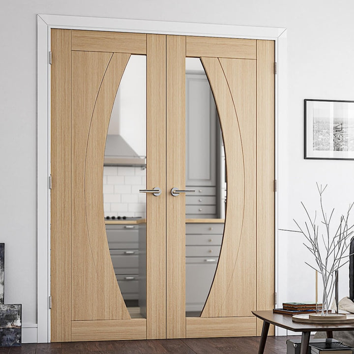 Ravello Oak Clear Glazed Door Pair -  The Ravello Oak door has a half circle design which when used as a pair of doors creates a vertical circular design, the door frame is primed white and surrounded by off white walls. The skirting and architrave are also white, flooring and the coffee table are in the picture are in a darker almost walnut colour  