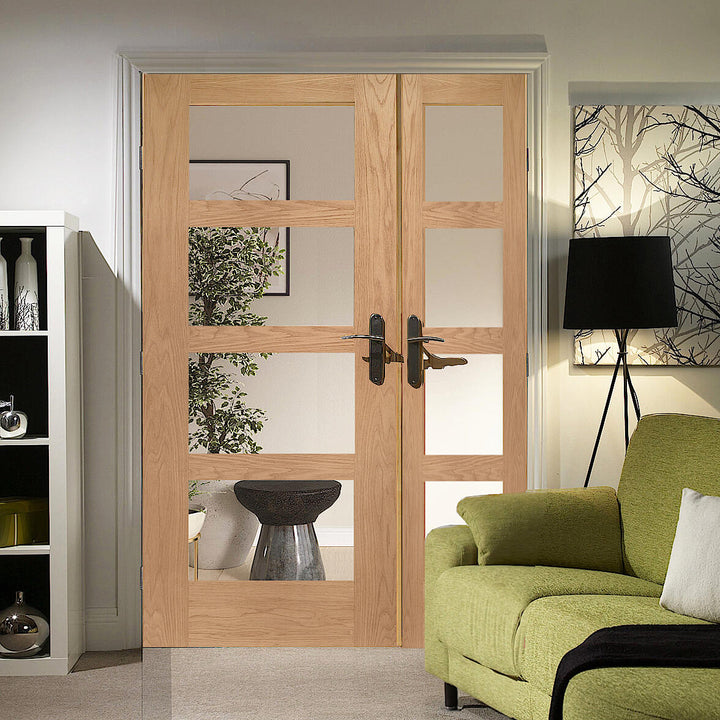 Oak Shaker 4 Light Offset French Doors With Demi Panel - Oak shaker style offset French doors with large single door and smaller door, these have traditional handles with a backplate on them, doors are fitted into a white frame and white architrave. There is a floor lamp to the right of the image and a green sofa can also be seen to the bottom left of the images.